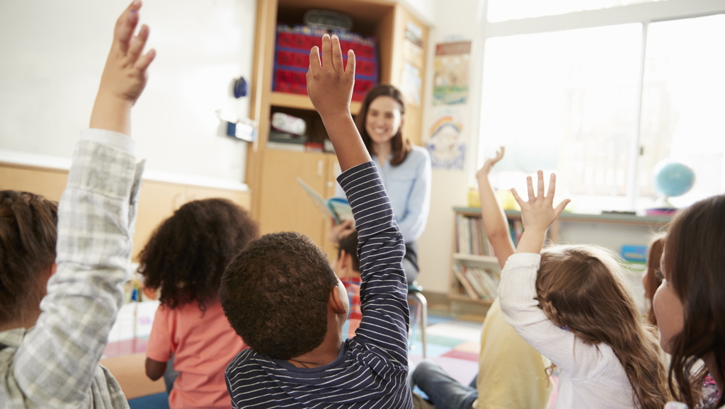 Children with hands raised to answer the teacher's question in a classroom